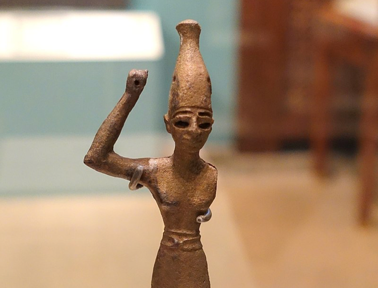 A bronze figure of a god wearing a tall, bowling-pin-shaped headdress, and holding aloft his right hand where a spear or tool existed at one time. 