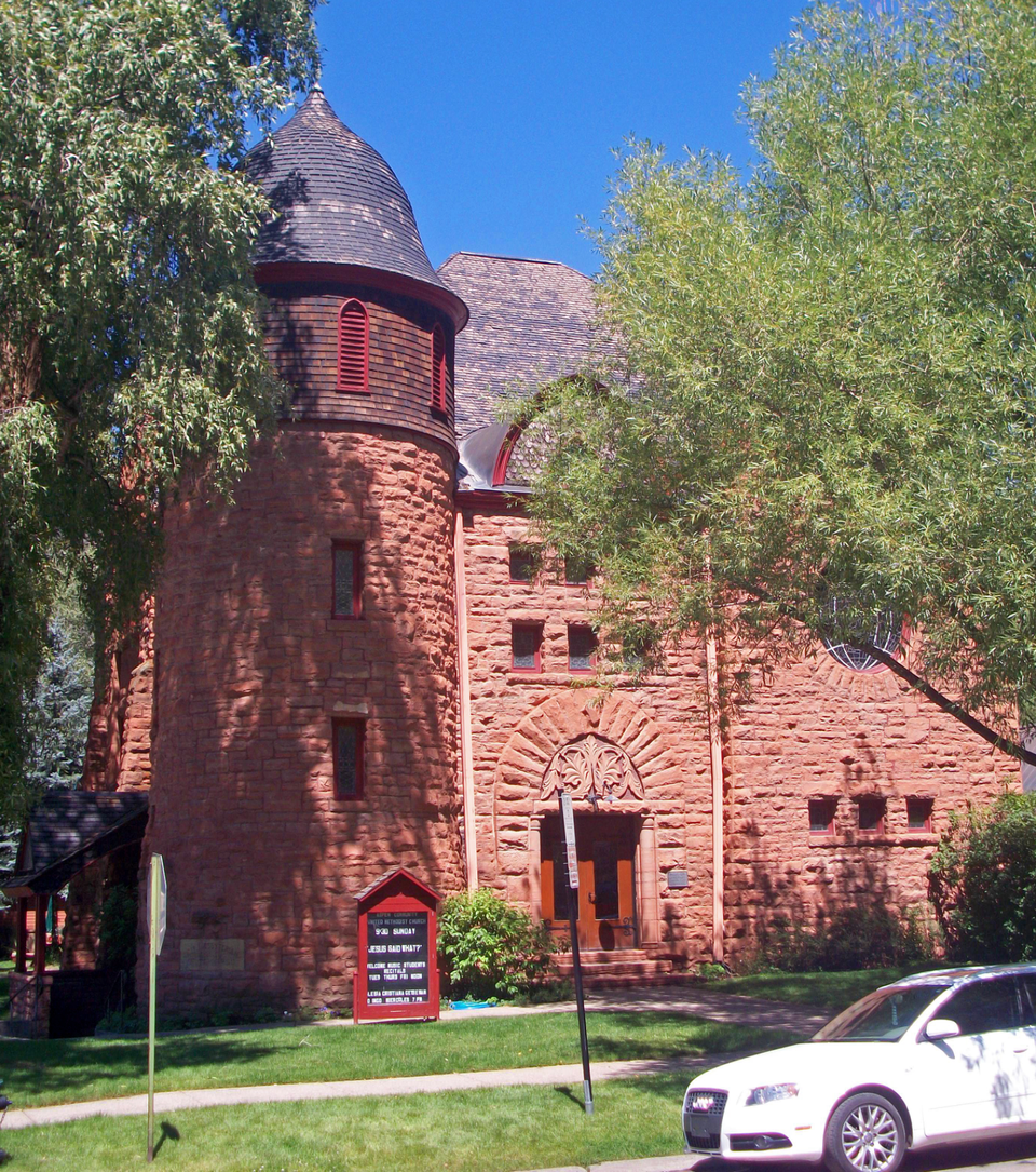 Bright sun shines on a red sandstone church of the late 19th or early 20th century. Green grass and shade trees adorn the front lawn. 
